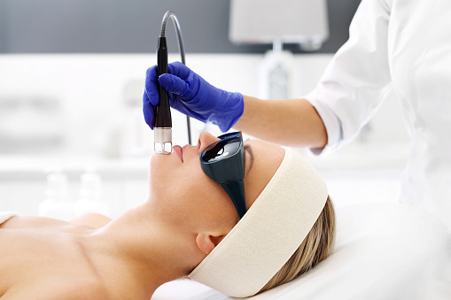 A woman relaxes while a beautician performs laser skin treatment on her face.