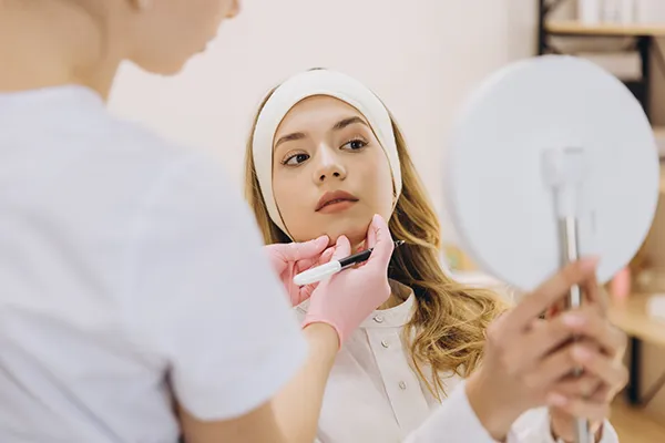 A woman inspects her face in the mirror as a dental assistant stands nearby, ready to mark the parts of her face that will receive dermal fillers.